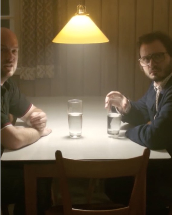 zwei Männer sitzen an einem Tisch über dem eine Lampe hängt und kegelförmig LLicht auf den Tisch wirft. Sie haben Wassergläser vor sich stehen. und blicken wütend in Richtung Kamera.